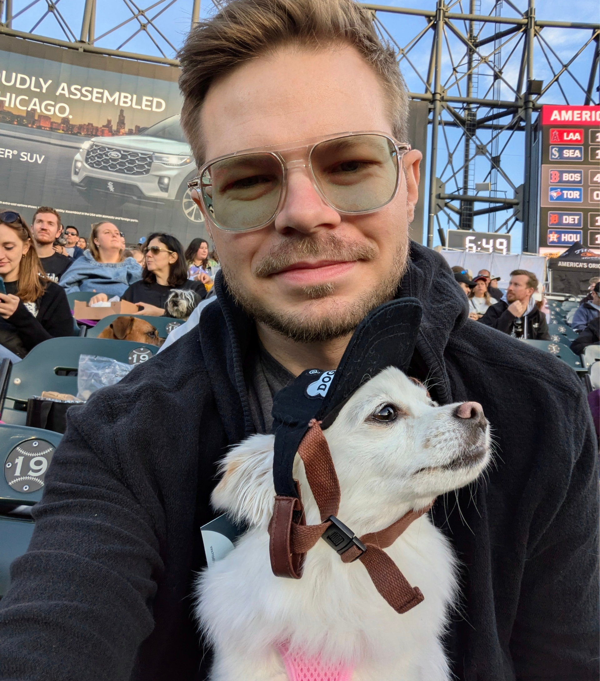 Greg holding a small white dog while in the stands at a baseball game.